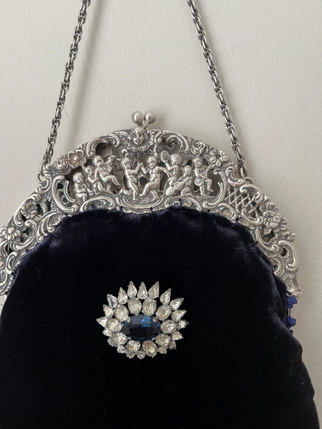 ②.1920年代银色框改造丝绒手袋（Peruzzi）；爱尔兰手艺人Cathy；古董银色手袋框，内侧刻有Peruzzi Boston Silver标记（因无925印猜测多为镀银或银色合金），饰以浮雕小天使（cherubs / putti）与繁复纹样。原始链条仍在。袋身以深蓝色丝绒制成，下缘配有黑色珠饰流苏；正面别有一枚可拆卸的捷克玻璃胸针。内衬为 Duchesse 丝绸，并以丝带与钩针花装饰。整体由古董框架与多种复古元素重构而成。银色金属/织物/玻璃等；尺寸约18×30cm（含流苏）；古董材质改造·孤独品